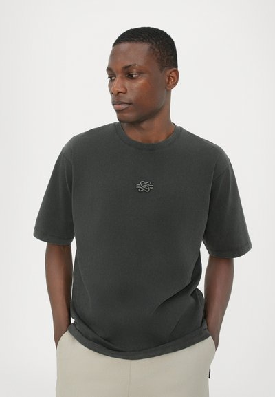 T-shirt gris foncé à manches courtes en coton avec un col rond et un logo texturé sur la poitrine, associé à un pantalon de couleur claire.