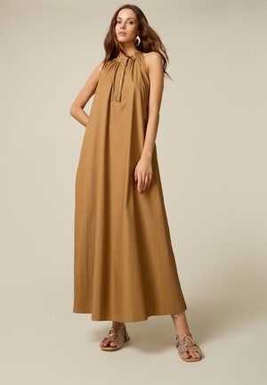 Kobieta ubrana w bezrękawną brązową sukienkę maxi z marszczonym dekoltem i plisą z złotymi guzikami, zestawioną z sandałami w wzór skóry węża.