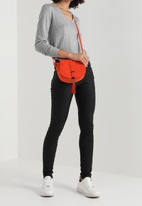 Červená crossbody kabelka s třásněmi, hladkým povrchem a nastavitelným popruhem. Nese se s šedým topem, černými kalhotami a bílými teniskami.