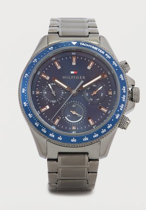 Montre en métal argenté avec une lunette tachymétrique bleue. Cadran sombre avec trois sous-cadrans, aiguilles argentées et dorées, et index des heures marqués.