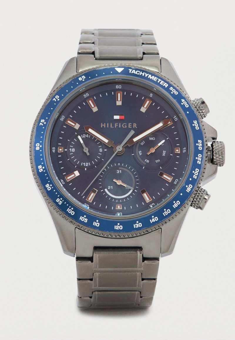 Orologio in metallo argentato con lunetta tachimetrica blu. Quadrante scuro con tre contatori secondari, lancette argento e oro e indici delle ore marcati.