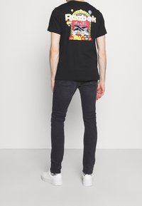 Černé bavlněné tričko s barevným potiskem na zádech, s logem a různými vzory. Kombinováno s tmavými džíny a bílými teniskami.