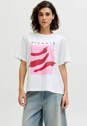 Kvinde med krøllet hår iført en hvid T-shirt med tre røde chilipebre og ordet "PICANTE", parret med lyseblå jeans.