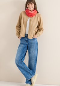 Jeune femme souriante en veste beige, écharpe rouge, jeans bleus et baskets blanches, debout les mains dans les poches contre un mur uni.