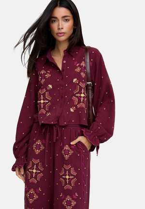 Frau trägt ein bordeauxrotes Hemd mit Knöpfen und passende Hose mit gelber und pinker geometrischer Stickerei, sie trägt eine dunkelbraune Umhängetasche.