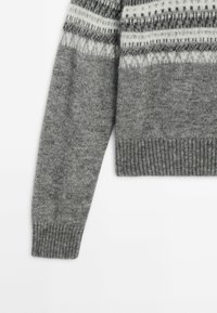 Manche de pull gris tricoté avec un poignet côtelé et un motif en blanc et gris foncé sur la partie supérieure.