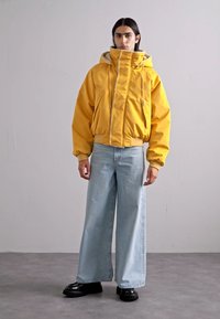 Yellow padded jacket with high collar, zip front, and drawstrings, paired with loose light blue jeans and black chunky boots.