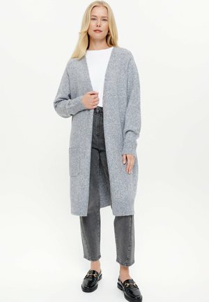 Grijze gebreide cardigan met open voorkant, geribbelde manchetten en zijzakken, gedragen over een wit shirt en gecombineerd met grijze jeans en zwarte instappers.
