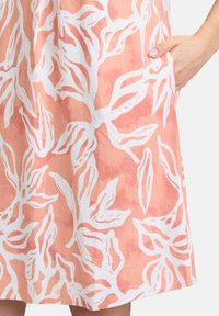 Ein helles Korallenkleid mit einem weißen Blattmuster, aus weichem Stoff, mit lockerer Passform und seitlichen Taschen.