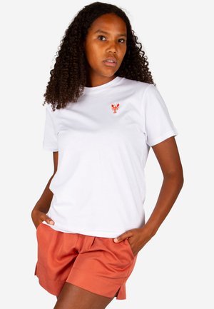 Woman with curly hair wearing a white T-shirt with a small orange lobster emblem and rust-colored shorts, hands in pockets.