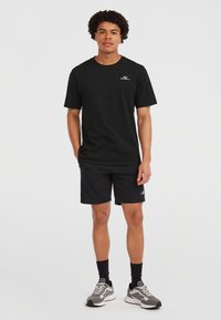 Black cotton T-shirt with a round neck and short sleeves, paired with black shorts. Notable logo on the chest, grey sneakers with a textured sole.