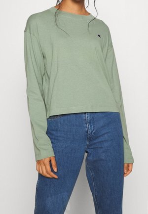 Osoba ubrana w luźną bluzkę na długi rękaw w kolorze szałwi zielonej, wpuszczoną w wysokowalne niebieskie dżinsy na jednolitym tle.