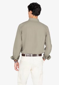 Camisa de manga larga de color verde oliva claro con un cuello y puños abotonados. Se lleva con pantalones color crema, mostrando un cinturón marrón con un emblema.