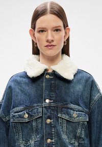 Τζάκετ από τζίν με λευκό ψεύτικο γούνινο γιακά, κλείσιμο με κουμπιά και δύο τσέπες στο στήθος. Το ύφασμα έχει μέτριο πλύσιμο με υφή.