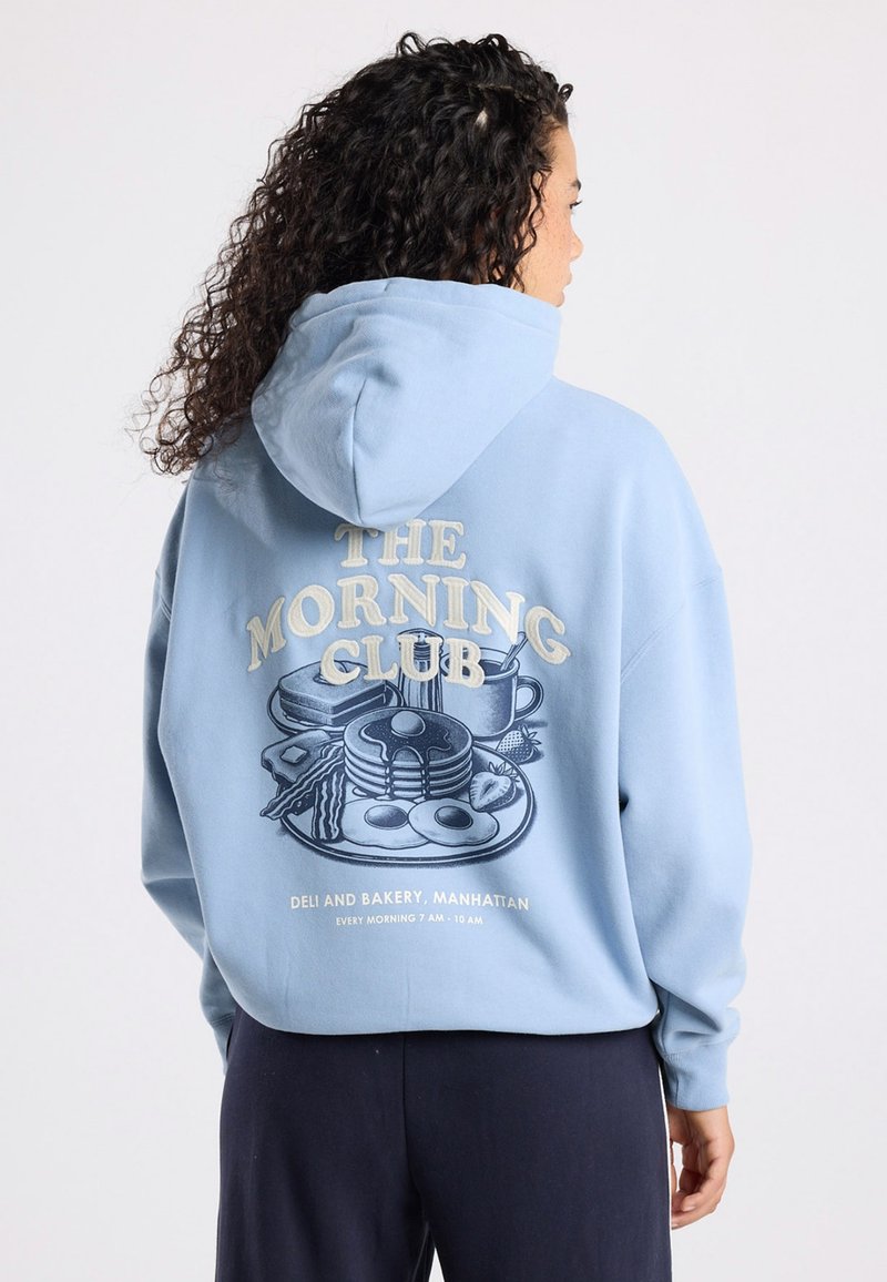 Hoodie bleu clair avec une impression graphique d'une scène de petit-déjeuner au dos. Comprend du texte et des illustrations d'articles alimentaires et d'une tasse.