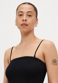 Μαύρη μπλούζα με σφικτές ραφές και λεπτές τιράντες, φτιαγμένη από υφή υφάσματος και εφαρμοστή σιλουέτα. Μικρό λουλουδάτο τατουάζ στο χέρι, ορατά σκουλαρίκια κρίκοι.