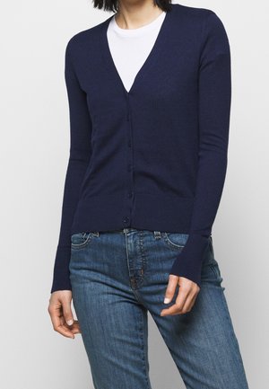 Slim-fit marineblauwe cardigan met knopen, gedragen over een wit overhemd, gecombineerd met blauw spijkerbroek met medium wassing.