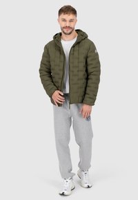 Olijfgroene gewatteerde jack met capuchon, met een ritssluiting aan de voorkant en getextureerde vierkanten. Draagt over een wit T-shirt en grijze joggingbroek.