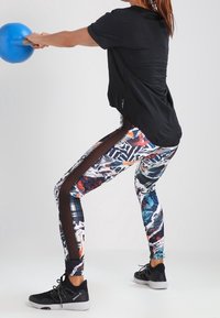 Persona con leggins de patrones coloridos y zapatillas negras moviendo una kettlebell azul con ambas manos.