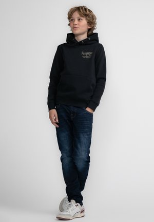 Felpa nera con cappuccio, tasca frontale e stampa del logo, abbinata a jeans blu scuro e sneakers bianche. Sfondo semplice.