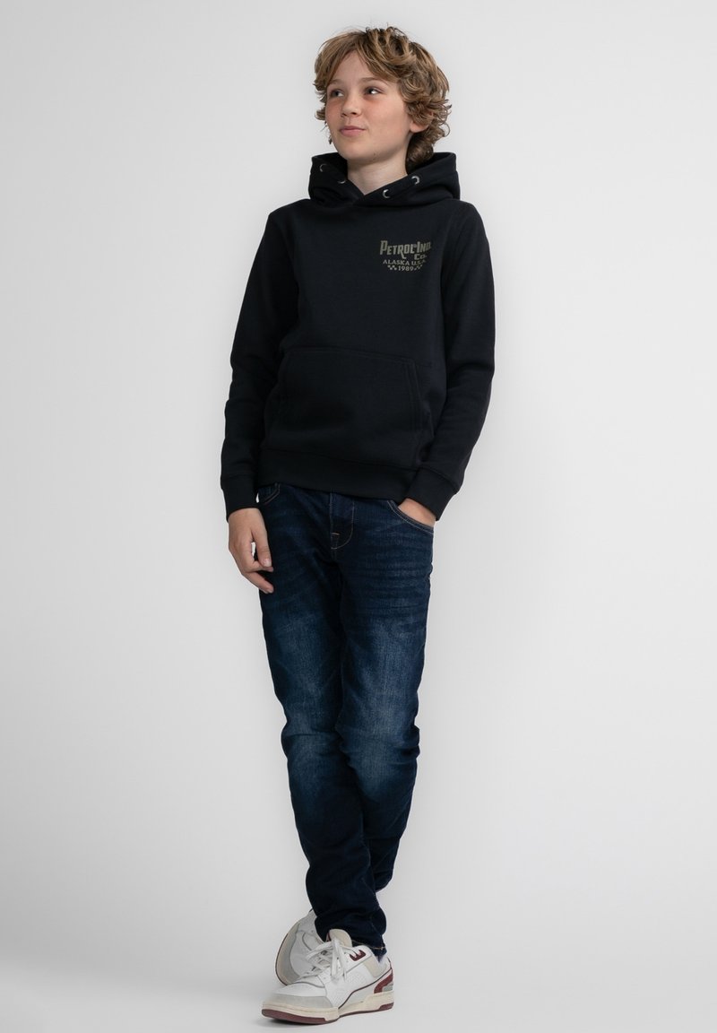 Felpa nera con cappuccio, tasca frontale e stampa del logo, abbinata a jeans blu scuro e sneakers bianche. Sfondo semplice.