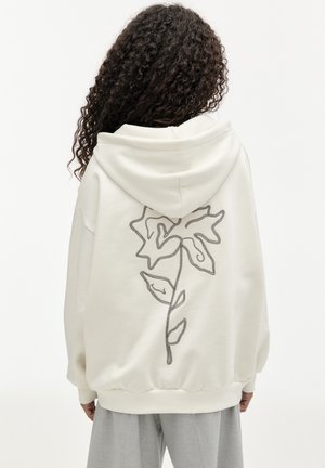 Personne aux cheveux bouclés portant un sweat à capuche blanc avec un grand motif floral simple en contour noir dans le dos et un pantalon gris clair.