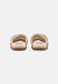 Par de zapatillas de pelaje en color beige con interior de piel de cordero, sin respaldo, y suelas de goma marrón, vistas desde el talón sobre un fondo blanco.