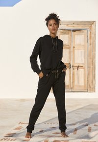 Schwarzes Hoodie- und Jogginganzug-Set aus weichem Stoff. Das Hoodie verfügt über einen Kordelzug mit goldenen Akzenten. Die Hose hat Taschen und ein gesticktes Detail.
