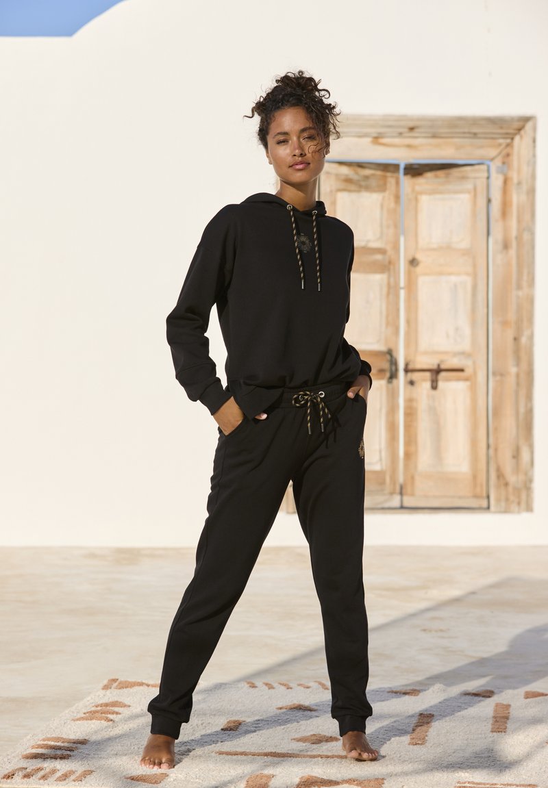 Schwarzes Hoodie- und Jogginganzug-Set aus weichem Stoff. Das Hoodie verfügt über einen Kordelzug mit goldenen Akzenten. Die Hose hat Taschen und ein gesticktes Detail.
