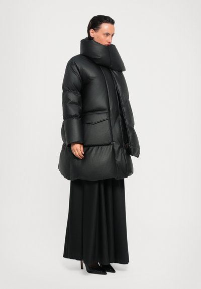 Πυκνό μαύρο παλτό με ψηλό λαιμό, τσέπη στο στήθος και πλαϊνές τσέπες. Λαμπερή υφή και σχεδίαση έως το γόνατο, συνδυασμένο με μια μακριά μαύρη φούστα.