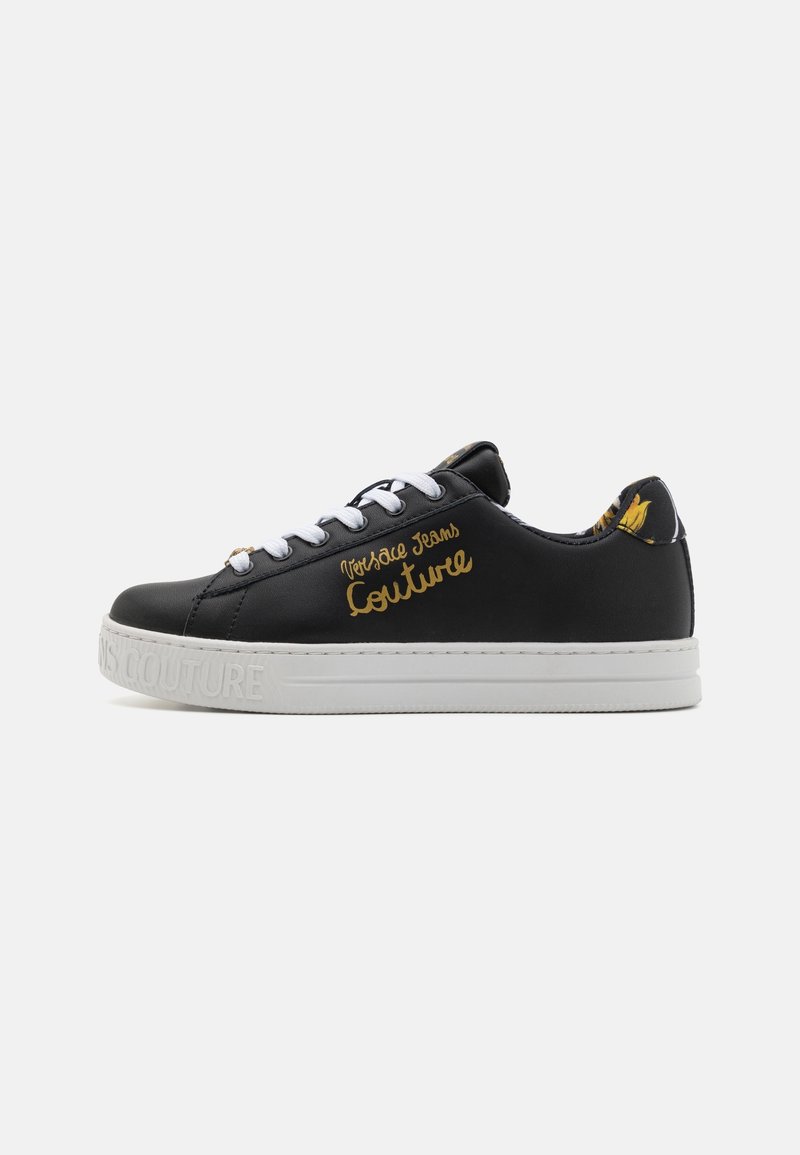Baskets basses noires avec semelle et lacets blancs, inscription dorée "Versace Jeans Couture" sur le côté, et motif floral sur la languette du talon.