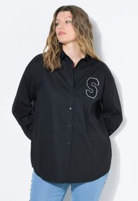 Camicia nera con bottoni in cotone, dotata di maniche lunghe, colletto a punta e una lettera "S" ricamata in bianco sul lato sinistro del petto.