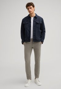 Giacca in denim navy con due tasche frontali, t-shirt bianca a girocollo, pantaloni tapered color beige chiaro e sneakers bianche. Look semplice e casual.