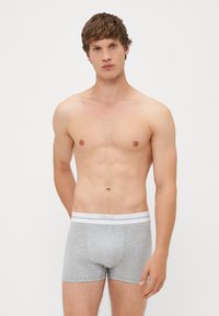 Boxeri gri din bumbac cu un tiv elastic alb. Tivul prezintă branding subtil. Modelul stă cu o expresie neutră.