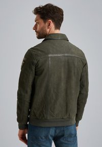 Chaqueta de cuero verde oliva con dobladillo y puños acanalados. Presenta un cuello puntiagudo, costuras sutiles y un parche bordado en el hombro.