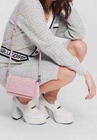 Różowa torebka zamszowa z logo, srebrne okucia, prostokątny kształt; zestawiona z białymi butami na platformie z złotym akcentem. Szary teksturowany strój.