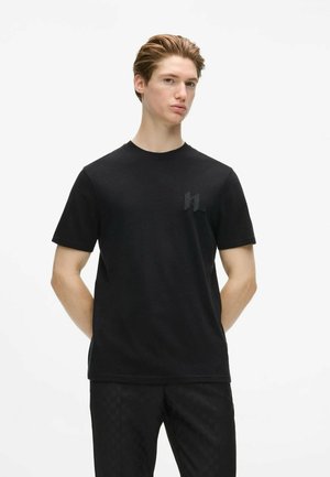 Jeune homme à la peau claire et aux cheveux coiffés portant un t-shirt noir uni à manches courtes et un pantalon noir, debout devant un fond blanc uni.