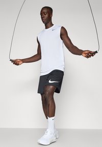 Biely bezrukávový športový dres s čiernym logom Nike, spárovaný s čiernymi šortkami s bielym logom Nike swoosh. Biely tenisky a ponožky.