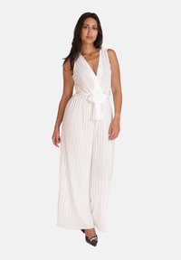 Hvid plisseret jumpsuit med dyb V-udskæring, en bindebælte i taljen og brede ben. Stylet med sorte spidse sko og minimale accenter.