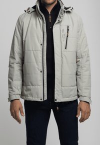 Chaqueta aislante gris claro con capucha, bolsillo en el pecho con cremallera y bolsillos laterales. Presenta un diseño acolchado y puños ajustables.