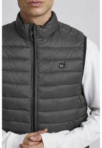 Chaleco de plumas gris con un cuello alto, que presenta un cierre de cremallera y un diseño acolchado horizontal. Acentos en negro y un pequeño logo en el pecho.