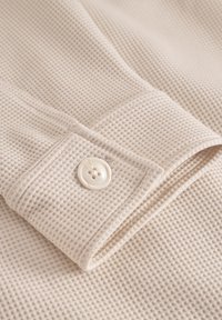 Beež tekstuuriga kangas, millel on vahvlipattern, koos volditud mansetiga, millel on ümmargune valge nupp ja õmmeldud servad.