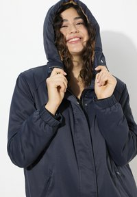 Marinblå vadderad jacka med huva, med elastiska ärmslut, dragkedjestängning och sidofickor. Tyget verkar slätt och vattenavvisande.