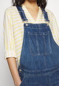 Denimoverall med justerbar silverdetaljer, en framficka och en mörkblå färg, kombinerat med en vit skjorta med gula ränder.