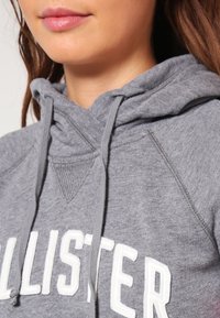 Sudadera con capucha gris de tela texturizada, con letras blancas en el frente y cordones en la capucha para ajuste regulable.