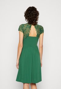 Zielona sukienka z koronkowym tyłem i krótkimi rękawami, z dopasowanym gorsetem i rozkloszowanym dołem. Gładka tekstura tkaniny z zaznaczoną talią.