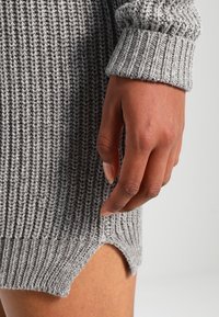 Grå stickad klänning med ribbad textur, som har en sidoklyfta och breda ärmslut. Den hand som visas vilar på klänningens söm nära fållen.