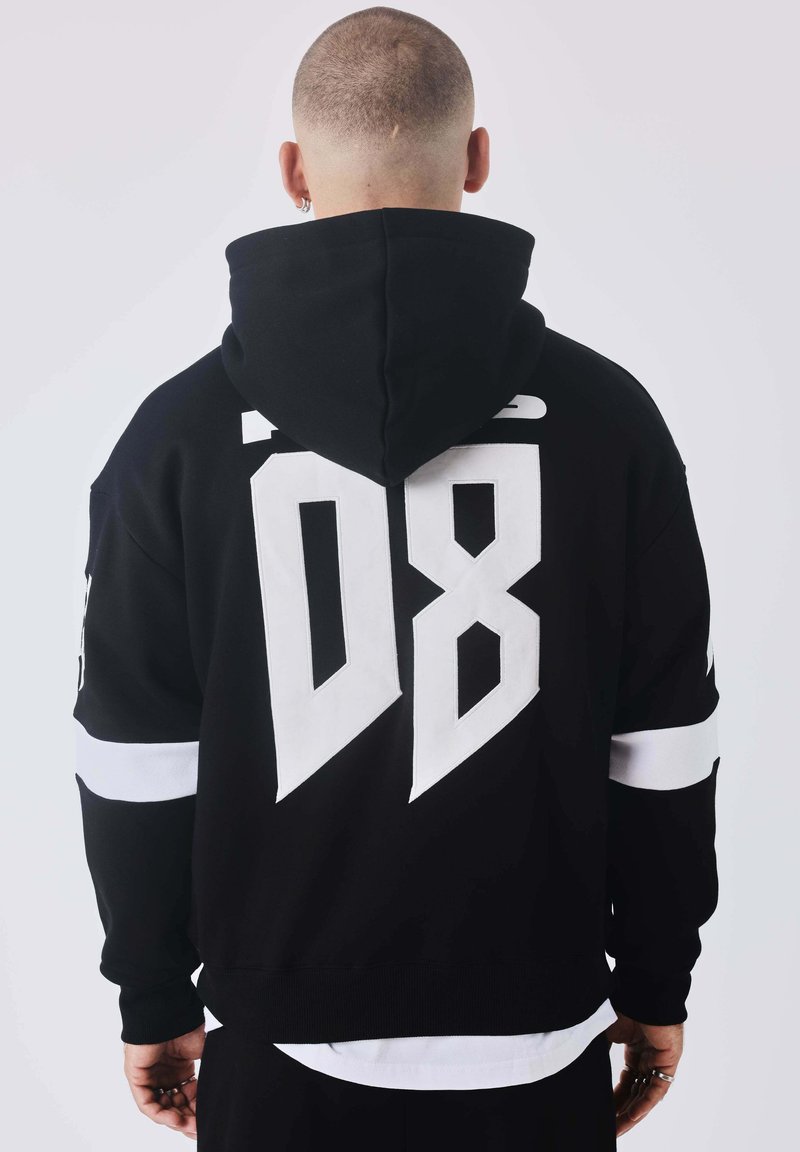 Personne au crâne rasé portant un sweat à capuche noir avec un grand « 08 » blanc dans le dos, debout devant un fond uni, dos tourné au spectateur.