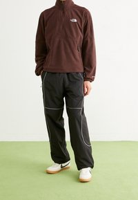 Pullover in pile marrone con zip a mezzo vano, pantaloni sportivi neri con piping bianco e sneakers bianche con suole in gomma.