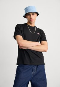Black cotton t-shirt with short sleeves and a small logo. Worn with blue denim jeans and a light blue bucket hat featuring text patterns.